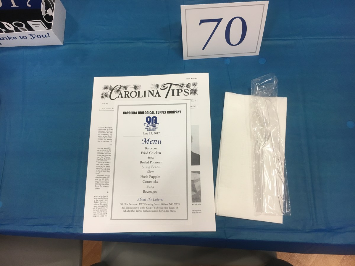 The table setting for a corporate luncheon including menu card, historical newsletter, table number, table centerpiece, napkin and disposable utensils